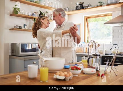 Liebe, Frühstück und ältere Paare tanzen in der Küche für Romantik und glückliches Leben zusammen im Haus. Verheiratete Rentner lächeln glücklich Stockfoto