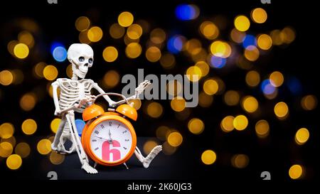 Ein menschliches Skelett winkt mit der Hand und begrüßt einen orangen Wecker auf schwarzem Hintergrund, Bokeh. Es ist Halloween-Zeit. Grußkarte Stockfoto