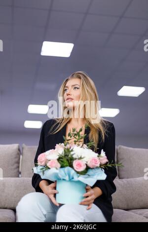 Porträt von attraktiven schönen und stilvollen jungen Frau mit Blumenstrauß in den Händen. Frau sitzt auf dem Sofa und hält Pappkarton wi Stockfoto