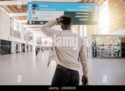 Rückansicht eines afroamerikanischen Geschäftsmannes, der alleine unterwegs ist und in einem Bahnhof steht, während er sich seine Reisezeiten ansieht und besorgt aussagt Stockfoto
