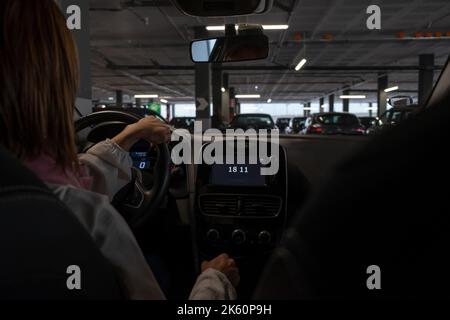 Eine Frau, die hinter dem Steuer auf einem überdachten Parkplatz sitzt. Ansicht vom Rücksitz in einem niedrigen Winkel zum Armaturenbrett des Fahrzeugs. Stockfoto