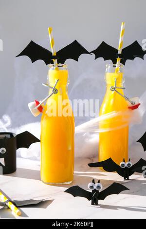 Halloween-Mocktails. Oranger alkoholfreier Cocktail für Halloween-Party auf weißem Tisch. Die Trinkhalme sind mit geschnittenen Papierfledermäusen verziert. Halloweens gruselig Stockfoto