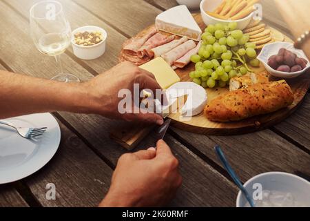 Unbekannter Mann, der Käse aus einer Vielzahl von Snacks auf einem Tapas-Holzbrett vor dem Haus nimmt und schneidet. Zum Mittagessen werden Brot, frische Trauben und Aufschnitt serviert Stockfoto