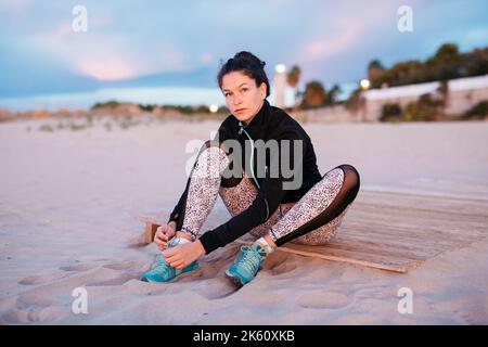 Junge Frau in Sportkleidung, die Schnürsenkel schnürt, bevor sie bei Sonnenuntergang am Strand arbeitet Stockfoto