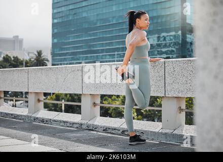 Eine junge Frau, die Trainingskleidung trägt und ihre Beine streckt, während sie auf einer Brücke steht und die Aussicht auf die Stadt betrachtet. Hispanischer Athlet Stockfoto