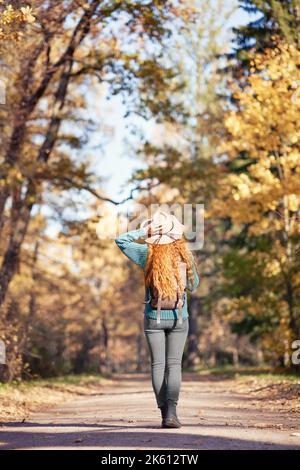 Eine junge rothaarige Frau geht durch den Herbstpark. Ein Mädchen in einem beigen Hut blickt auf den Herbstwald Stockfoto