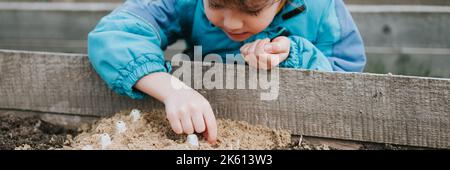 Frühling Pflanzung Aussaat im Bauerngarten. Kleine sechs Jahre alte Kind Junge Landwirt Gärtner Pflanzen und säen Gemüsesamen im Boden im Bett. Gartenarbeit und beginnen Stockfoto