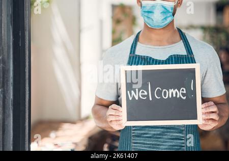 Nahaufnahme eines Mannes mit gemischter Rasse, der eine Gesichtsmaske trägt und eine Tafel mit dem Wort Willkommen in einem Café oder Geschäft hält. Hände eines Mannes, der hält Stockfoto