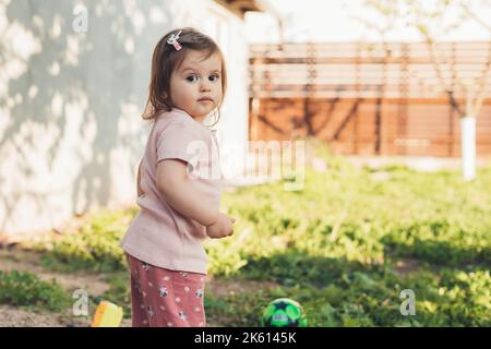 Porträt eines entzückenden kaukasischen Mädchens, das auf die Kamera schaut, während es seine Großeltern im Dorf besucht und fröhlich im Garten spielt Stockfoto