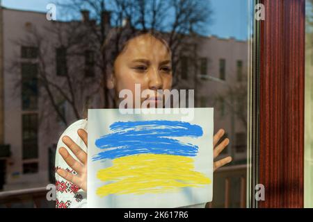 Ein Mädchen in einem gestickten Kleid blickt aus dem Fenster, hält ein weißes Blatt mit einem Bild und einer Inschrift. Das Mädchen protestiert gegen den Krieg.Ukraine Stockfoto