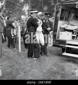 1960s, historisch, draußen bei einer County Show, helfen zwei uniformierte Mitglieder des St. john Krankenwagens einer verzweifelten Dame in den Rücken eines Notfahrzeugs, Buckinghamshire, England, Großbritannien. Die Geschichte von St. John Ambulance, Großbritanniens führender erste-Hilfe-Organisation, die seit über 100 Jahren auf Veranstaltungen aktiv ist, reicht bis ins 11.. Jahrhundert zurück, als die ersten Ritter von St. john in Jersusalem ein Krankenhaus für kranke Pilger aufbauten. Stockfoto