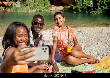 Ein großer Winkel glücklicher multiethnischer Freunde, die während der Sommerreise Selfie-Portraits auf dem Handy machen, während sie am Ufer des Sees sitzen Stockfoto