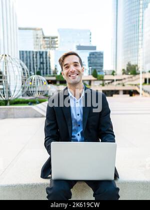 Positiver Kerl in elegantem Anzug sitzt auf der Bank in der modernen Stadt und browsing Computer während der Arbeit an Projekt im Sonnenlicht Stockfoto