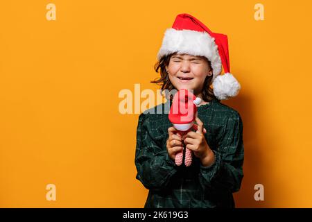Glückliches Mädchen in kariertem Kleid und Weihnachtsmann Hut greifen Spielzeug und Augen schließen, während für Weihnachtsgeschenk vor gelbem Hintergrund wünschen Stockfoto