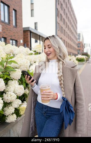 Blonde Frau in stilvoller Oberbekleidung stöbert im Frühjahr beim Mitnehmen auf der Straße der Stadt Stockfoto