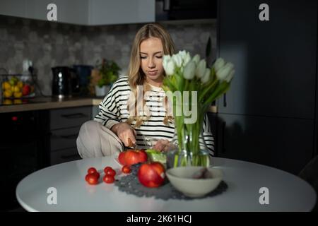 Charmante Blondine sitzt am Tisch in ihrer Küche, frisches Gemüse und Obst stehen vor ihr auf dem Tisch. Diät. Stockfoto