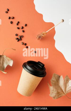 Kaffeebecher mit Naturkorkband. Kaffeetasse auf orange-weißem Herbsthintergrund mit trockenen Platanenblättern. Kaffeebohnen und Zuckerstick. Vertikal Stockfoto