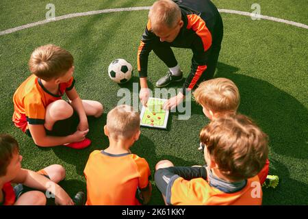 Fußballtraining. Fußballtrainer erklärt Spielregeln und Strategie mit Tablet, Karte. Sport Junior Team sitzt auf Rasen mit Trainer Stockfoto