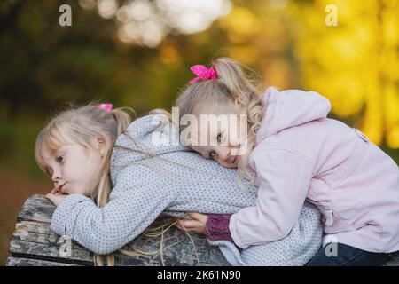 Zwei niedliche kleine Mädchen liegt auf einem gefallenen Baumstamm im Park und lächelt. Porträt eines lächelnden Mädchens, das auf dem Rücken ihrer Schwester liegt. Stockfoto