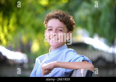 Junge glücklich Kind Junge entspannen auf der Bank im Sommer Park sitzen. Positives Kind, das die Sommerzeit im Freien genießt. Konzept für das Wohlbefinden von Kindern Stockfoto