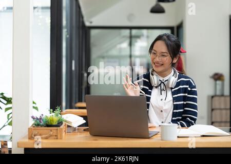 Lächelnde asiatische Geschäftsfrau im Headset winkende Hand, mit Laptop, Blick auf den Bildschirm, Schüler lernen Sprache, Mentor Lehrer Ausbildung online. Stockfoto