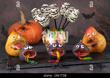 Gruselige Birnen- und Feigenmonster, Mama geformte Schokoladenkuchen-Pops und Gummiwürmer für Halloween-Party, Halloween Obst und Süßigkeiten Servieridee Stockfoto