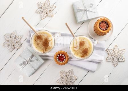 Weihnachtliche flache Anordnung von traditionellen Getränken und Gebäck auf weißem Holztisch. Gläser mit Eierkarton auf gestreifter Serviette, Kekse, Geschenkschachteln, dez Stockfoto