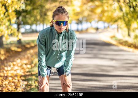 Müde junge Frau ruht sich nach einem Lauf mit einer intelligenten Uhr und Ohrhörern aus. Stockfoto