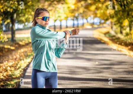 Eine junge Frau streckt an einem Herbsttag im Freien den Arm, trägt Uhren, Sonnenschutz und Kopfhörer. Stockfoto