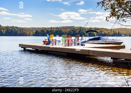 Motorboot an einem sonnigen Herbsttag an einem hölzernen Pier am Ufer eines Sees befestigt. Am Pier befinden sich farbenfrohe Sessel. Stockfoto