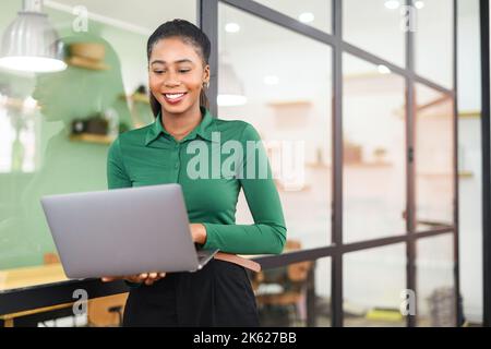 Eine intelligente afroamerikanische Geschäftsfrau trägt ein schickes Casual-Shirt und nutzt einen Laptop in Innenräumen, eine Büroangestellte hält einen Computer und schreibt eine Nachricht, lächelt und genießt ihre Arbeit Stockfoto