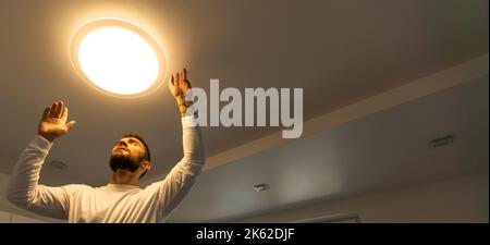 Ein junger Mann steht auf einer Stube und installiert einen großen Kronleuchter an der Decke Stockfoto