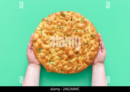 Die Hände der Frau halten eine frisch gebackene Rosmarin-Focaccia über einem grünen Tisch. Draufsicht mit hausgemachter Focaccia mit einer köstlichen Kruste. Stockfoto