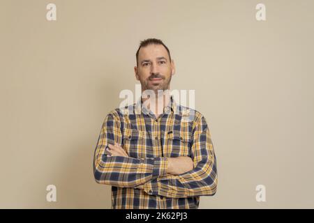 Fröhlicher, selbstbewusster kaukasischer Mann im karierten Hemd mit gekreuzten Armen auf der Brust, isoliert auf beigem Hintergrund. Moderner Geschäftsmann, der sich trifft Stockfoto