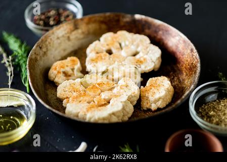 Blumenkohlsteak Kochen. Roher Blumenkohl, mit Gewürzen bestreut, liegt in einer Bratpfanne. Olivenöl, Kräuter, verschiedene Gewürze in der Nähe. Dunkler Hintergrund. Stockfoto