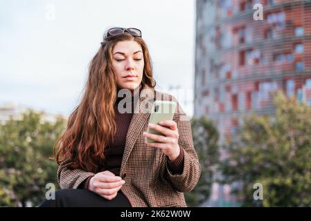 Niedriger Winkel der Frau in stilvollen karierten Mantel und Rollkragen Surfen Social Media auf dem Handy, während auf verschwommenem Hintergrund der Straße in der Stadt sitzen. Stockfoto