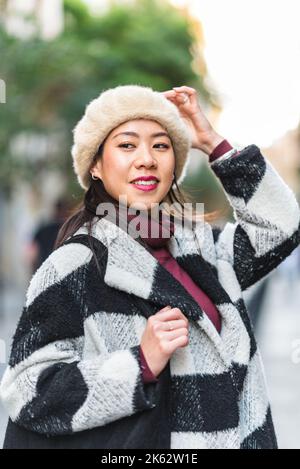 Inhalt Chinesisches Weibchen in stilvollem, warmem Mantel und Baskenmütze, das mit der Hand auf dem Kopf wegschaut, während es vor verschwommenem Hintergrund auf der Straße steht Stockfoto