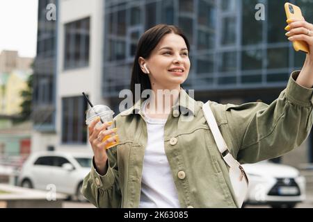 Glückliche junge Frau, die sich mit einem Cocktailgetränk in der Hand mit ihrem Mobiltelefon fotografiert, kaukasische Frau, die Selfie mit dem Smartphone auf der Straße macht. Stockfoto