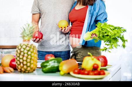 Junges Paar mit einer Schwangeren beim Kochen in der Küche mit gesundem vegetarischem Essen und Obst - Liebeskonzept mit Menschen zu Hause, die veganes Mittagessen zubereiten Stockfoto