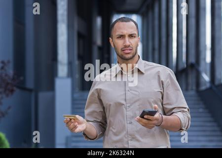 Porträt eines unzufriedenen und getäuschten Mannes, eines Geschäftsmannes vor dem Bürogebäude, der die Kamera ansah und die Bankkreditkarte und das Smartphone hielt. Stockfoto