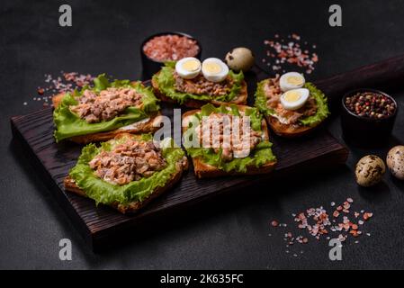 Köstliche frische Sandwiches mit Toast, Lachskonserven, Salat und Wachteleiern auf dunklem Beton Stockfoto