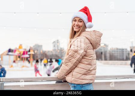 Outdoor-Porträt junge schöne modische Frau trägt Santa Hut, posiert Straße der europäischen Stadt. Winter Weihnachtsferien Konzept. Eislaufbahn Hintergrund. Lehnen Sie sich auf einen Zaun in der Nähe der Eislaufbahn Stockfoto