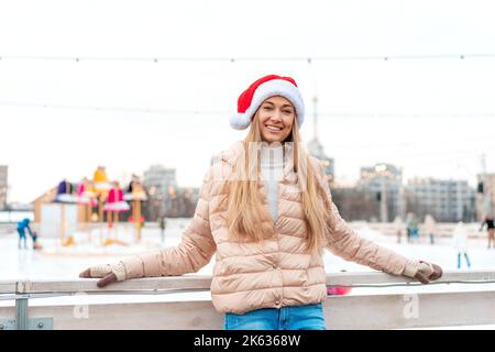 Outdoor-Porträt junge schöne modische Frau trägt Santa Hut, posiert Straße der europäischen Stadt. Winter Weihnachtsferien Konzept. Eislaufbahn Hintergrund. Lehnen Sie sich auf einen Zaun in der Nähe der Eislaufbahn Stockfoto