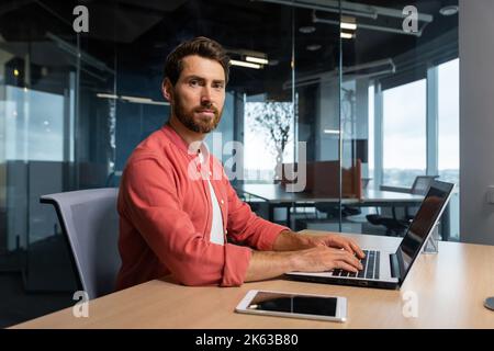 Portrait von reifen Programmierer Teamleiter Mann mit Bart und in rotem Hemd konzentriert und denkt Blick auf Kamera, Entwickler arbeiten innerhalb der modernen Firma mit Laptop für die Codierung. Stockfoto
