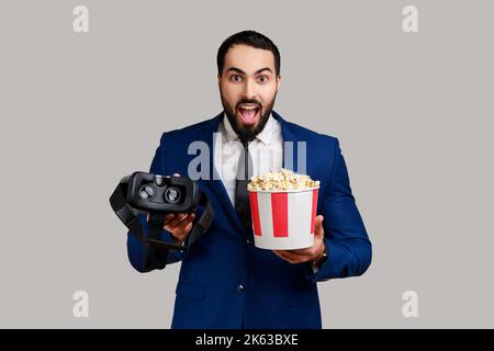 Überraschender bärtiger Mann, der ein VR-Headset in den Händen hält und Augmented-Reality-Brillen verwendet, die sich in der virtuellen Realität befinden und einen Film mit Popcorn ansehen. Innenaufnahme des Studios isoliert auf grauem Hintergrund. Stockfoto