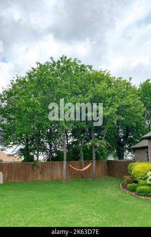 Außenaufnahme des Sommergartens mit hängender Hängematte zur Entspannung, Hängematte unter Bäumen im Hinterhof mit dem Zaun und dem Haus im Hintergrund. Entspannungskonzept. Stockfoto