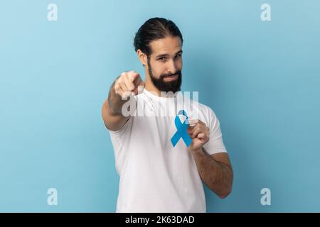 Porträt eines verantwortungsbewussten, gutaussehenden Mannes mit weißem T-Shirt und blauem Bewusstsein, Krankheitssymbol, zeigt auf Kamera, Unterstützung. Innenaufnahme des Studios isoliert auf blauem Hintergrund. Stockfoto