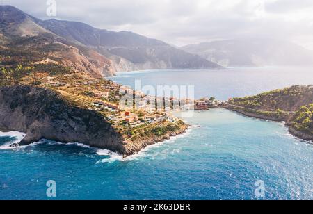 Malerische Luftaufnahme von bunten Fischerhäusern in Asos auf der Insel Cefalonia, Griechenland. Steile Steilhänge, die von blauen Wellen des Ionischen Meeres gespült werden. Stockfoto