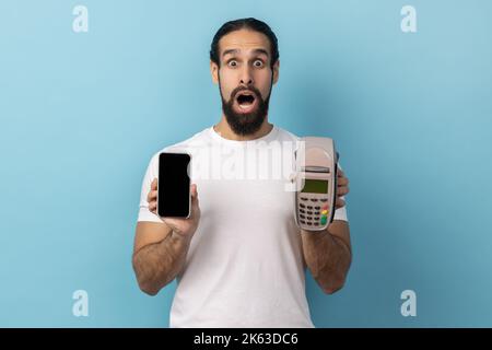 Porträt eines schockierten glücklichen Mannes mit Bart trägt weißes T-Shirt mit POS-Terminal und Handy mit leerem Display für Werbung. Innenaufnahme des Studios isoliert auf blauem Hintergrund. Stockfoto