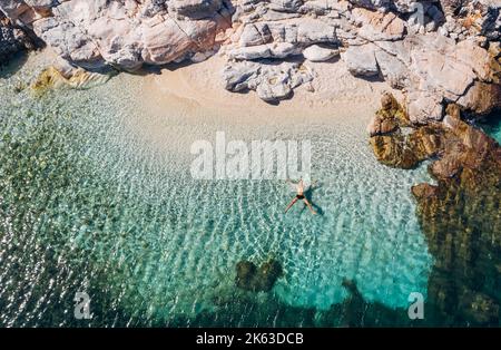 Luftaufnahme eines Menschen, der in STERNPOSE am felsigen Kiesstrand und in Sonnenbräunen auf dem Wasser liegt. Weiche Wellen waschen seinen Körper auf einer einsamen, einsamen griechischen Insel Stockfoto